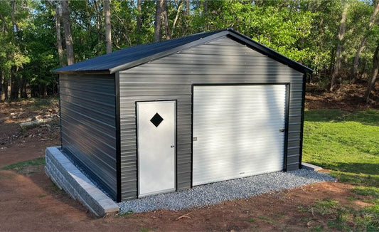 Metal storage shed with a diamond-shaped window and garage door in a wooded area.