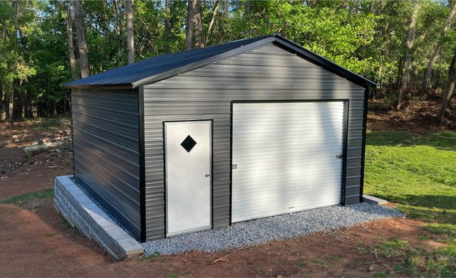 Metal storage shed with a diamond-shaped window and garage door in a wooded area.